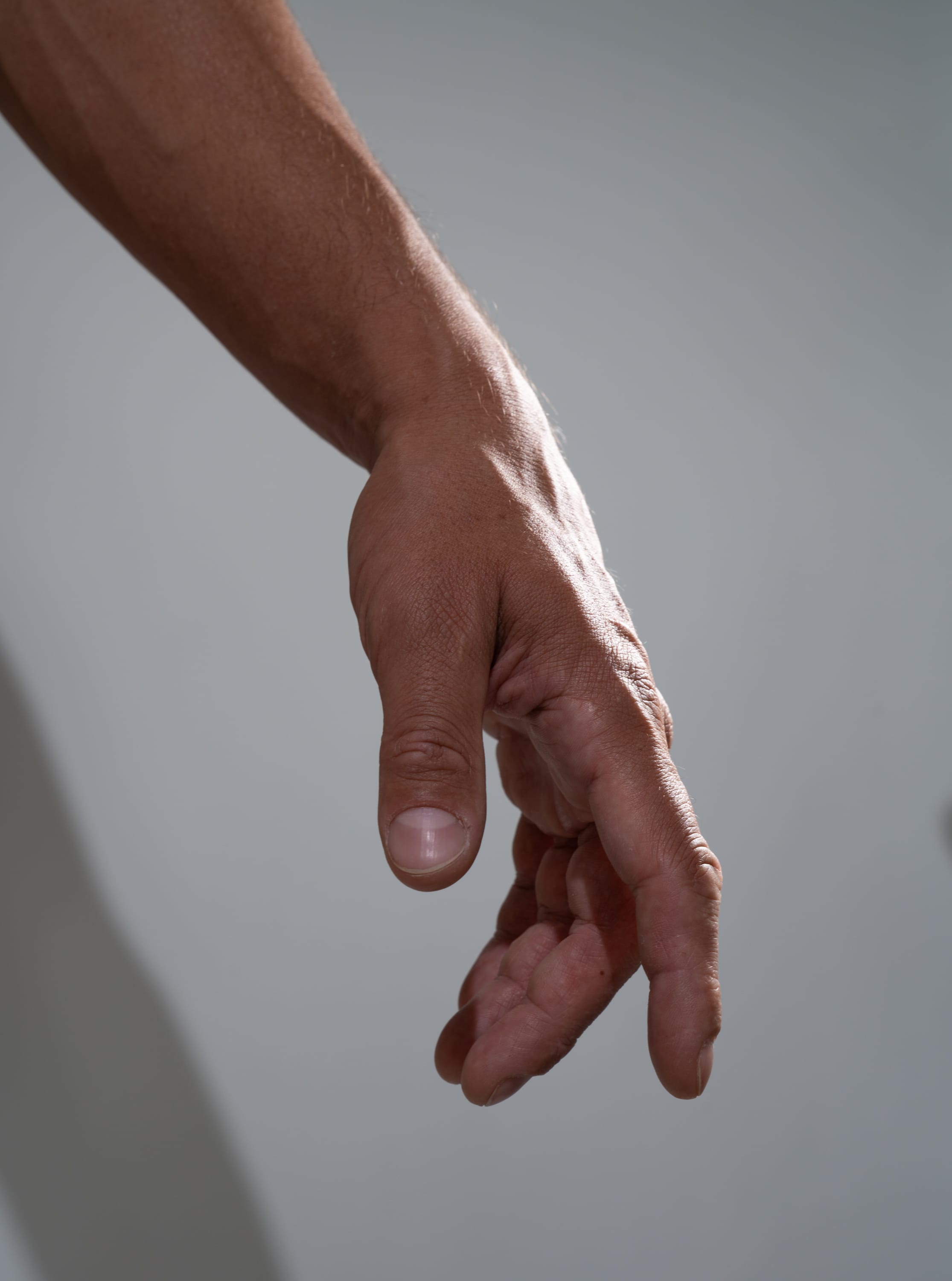 a closeup shot of a man's arm and hand extended downwards against a light gray background. the hand appears relaxed with slightly bent fingers. the skin tone is tan with hints of brown. the lighting is soft and likely natural, creating a gentle shadow on the wall behind the arm.