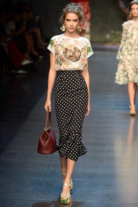 a runway model walks the dolce & gabbana fashion show. she's wearing a white floralprint blouse, a black and white polkadot pencil skirt, and highheeled shoes. she carries a burgundy handbag and has her hair styled in an updo.  another model in a floral dress is visible in the background. the overall feel is elegant and feminine, representing high fashion for a spring/summer collection.