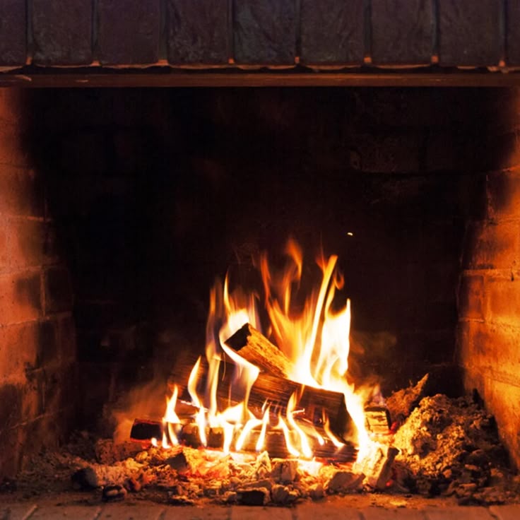 the image shows a fireplace with a fire burning. the fire is built with logs and has bright orange and yellow flames. the fireplace is made of brick and there is ash and embers at the bottom. the scene appears warm and cozy.