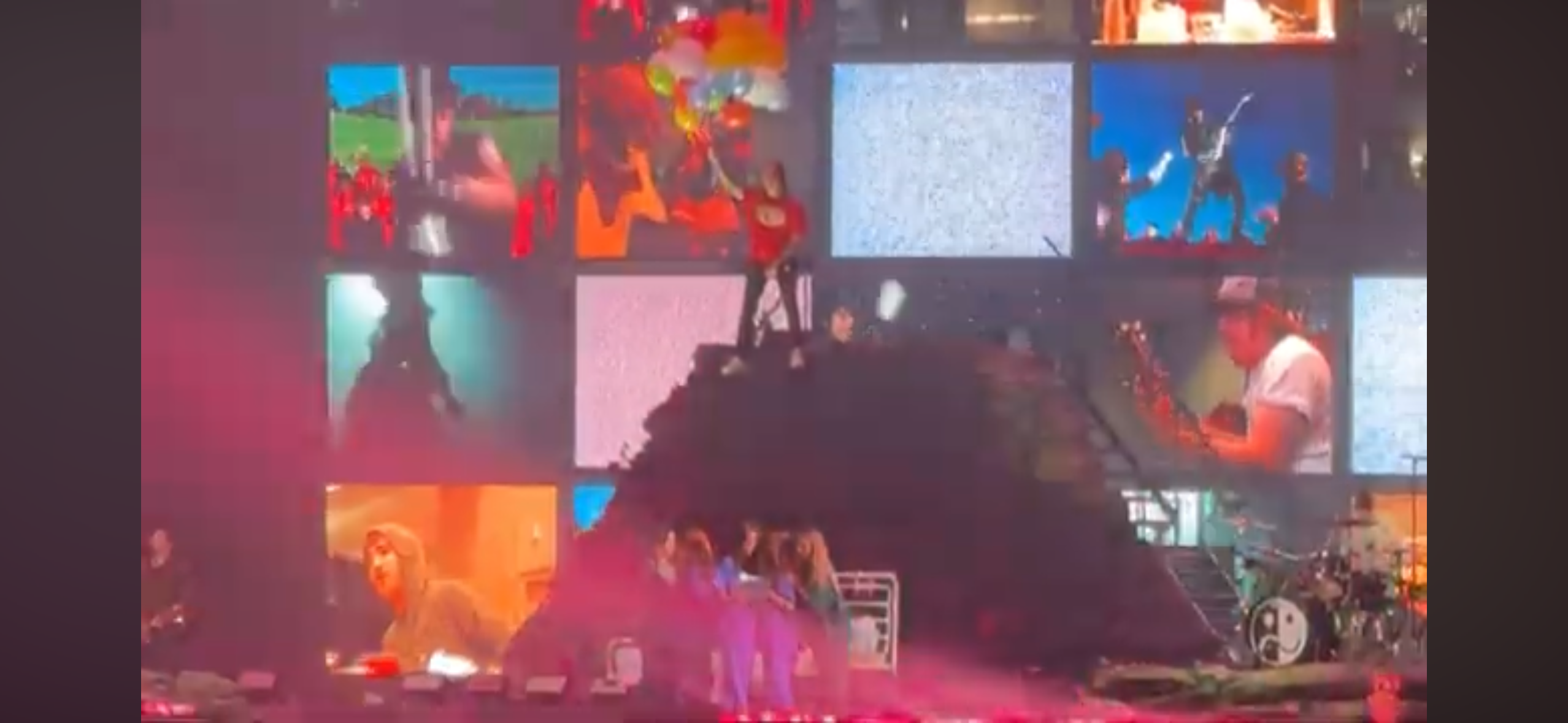 a rock band is performing on stage at a concert. the stage is lit up with bright lights and there are several video screens behind the band. the band is playing loud rock music and the crowd is cheering. the band is wearing casual clothes. there are many other musicians on stage. the concert is taking place in a large venue.
