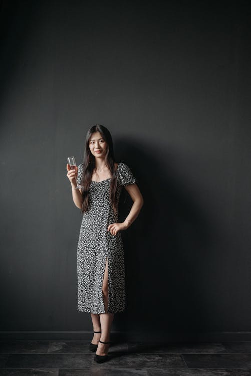 a full shot depicts a woman standing in front of a black background. she is wearing a floral print dress, and black heels. in her right hand she holds a wine glass.