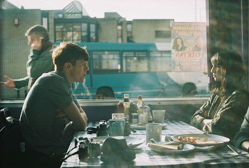 the image shows a cafe scene. two people, a man and a woman, are seated at a table near a large window. the man is looking down and the woman is looking out the window.  the setting appears to be an urban cafe, with a view of a city street and a bus outside. the table is set with breakfast food, cups of coffee and mugs. the light suggests it's daytime and sunlight is streaming in through the window. the overall mood is pensive and quiet. the image's aesthetic is reminiscent of vintage film photography with a slightly desaturated color palette.
