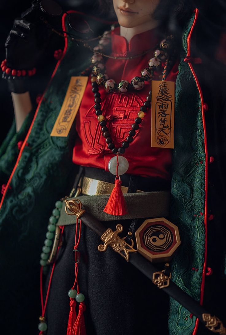 a closeup portrait of a doll dressed in a traditional chinese costume, highlighting intricate details. the doll is wearing a red, embroidered shirt, a green, embroidered robe with gold accents, and a gold belt. the doll has a jade necklace with black beads, and a sword with a golden handle and tassel. the doll's right hand is raised to its face, as if it is thinking or pondering. the overall look is dramatic and elegant, showcasing the beauty of traditional chinese fashion. the doll's expression is serene and enigmatic, adding to the overall mysterious and fascinating feel of the image.