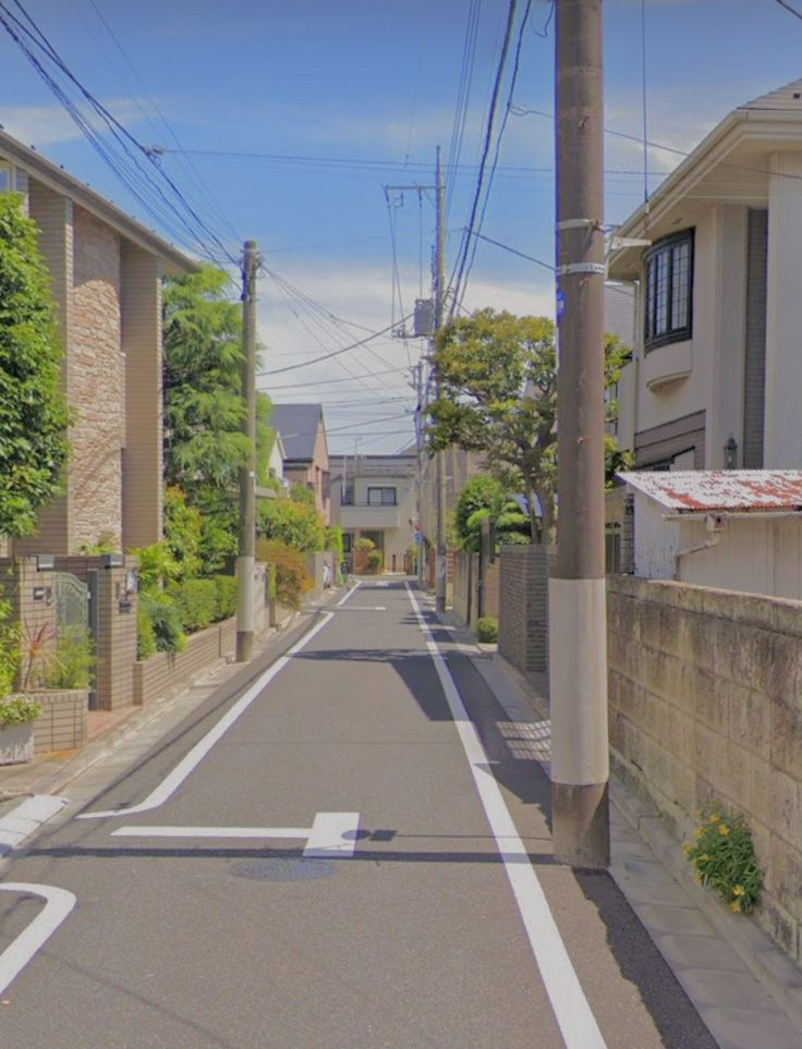 a street view of a residential neighborhood on a sunny day. the street is lined with houses and plants, and a utility pole stands on the right side of the road. the sky is blue with minimal clouds. power lines are strung across the sky. sidewalks are present on either side of the street.