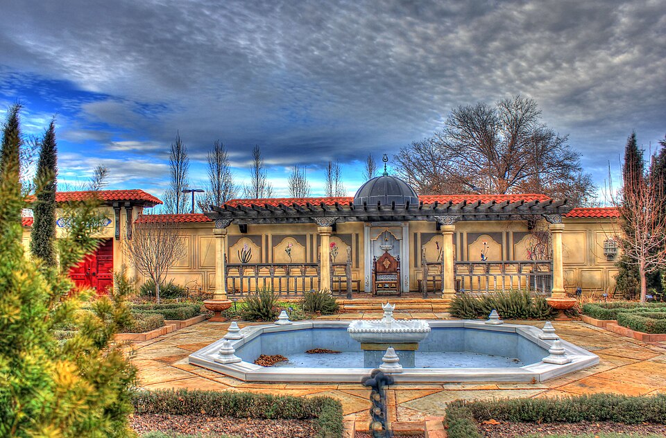 the image shows a garden with a fountain in the foreground and a gazebo in the background. the garden is wellmaintained with manicured hedges and trees. the gazebo has a pergola and a dome on top. the sky is cloudy. the architecture is middle easterninspired.