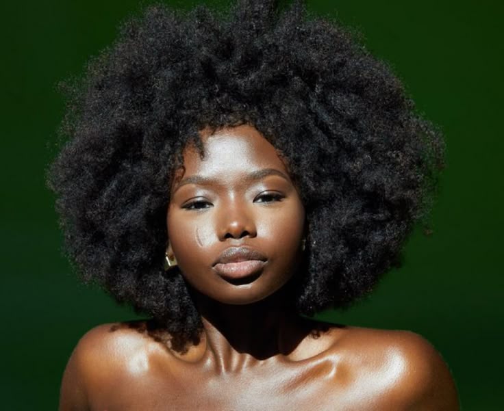 a portrait of a beautiful black woman with a large, natural afro hairstyle. she has dark skin and is wearing gold earrings. her expression is serious and confident. the background is a solid green color. her skin has a noticeable sheen, possibly due to lighting or product.