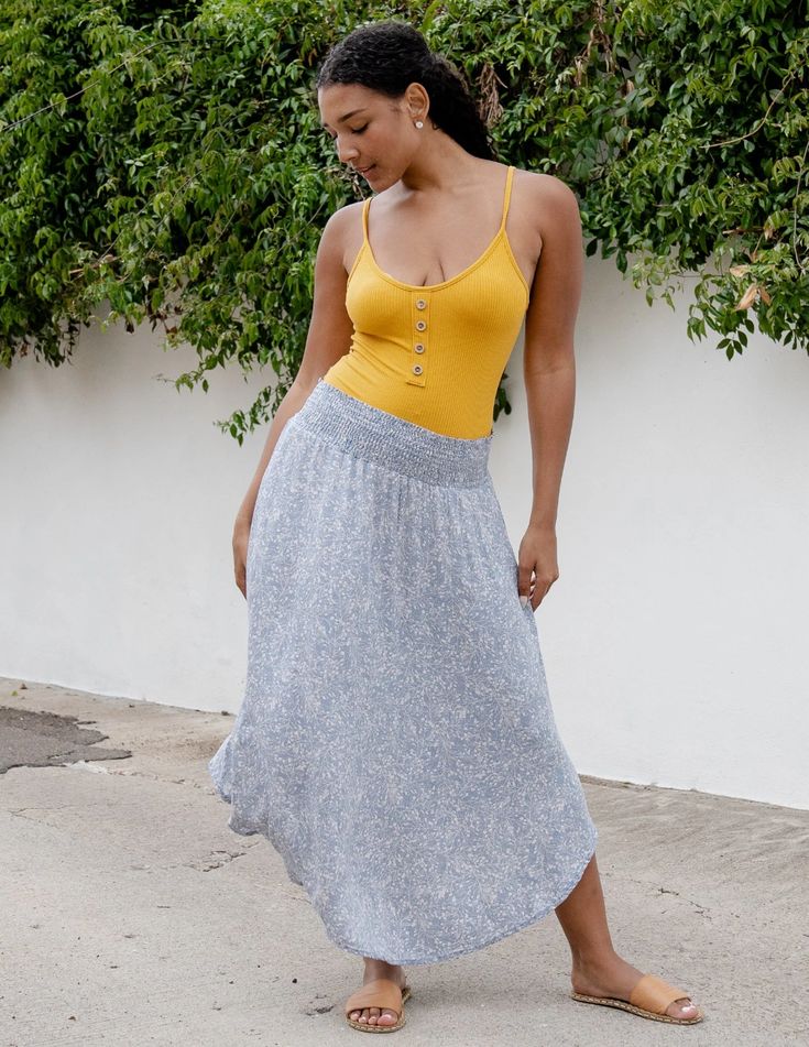 a woman with long dark hair is standing on a paved street in front of a white wall covered with green plants. she's wearing a mustard yellow tank top and a long, flowy blue and white floral skirt. she is also wearing brown sandals. she is posing with her head tilted down and her right leg slightly bent. the image is taken from eye level in natural, sunny lighting.