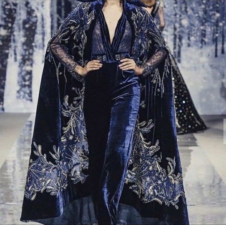 the image shows a model on a runway wearing a stunning navy blue velvet jumpsuit paired with an equally impressive floorlength cape. the cape is richly embellished with silver embroidery featuring intricate, swirling floral and possibly celestialinspired designs. the jumpsuit is sleek and formfitting, contrasting with the flowing, dramatic silhouette of the cape. the overall aesthetic is luxurious, elegant, and evocative of winter or a formal evening event.  the background is blurred but suggests a fashion show setting.