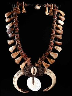 a necklace crafted from bone, teeth, and shell elements is displayed against a black backdrop. the necklace features numerous small bone or teeth pieces strung together, interspersed with dark wooden beads. a larger decorative element at the bottom comprises two tusklike bones flanking a textured rectangular piece with a shell attached. a white circular shelllike disc hangs below.