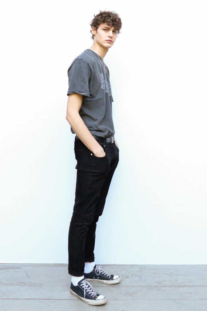 a full shot of a young man standing in front of a white background. he is wearing a gray tshirt, black jeans, and black sneakers with white socks. he has curly hair. he is standing with his hands in his pockets.