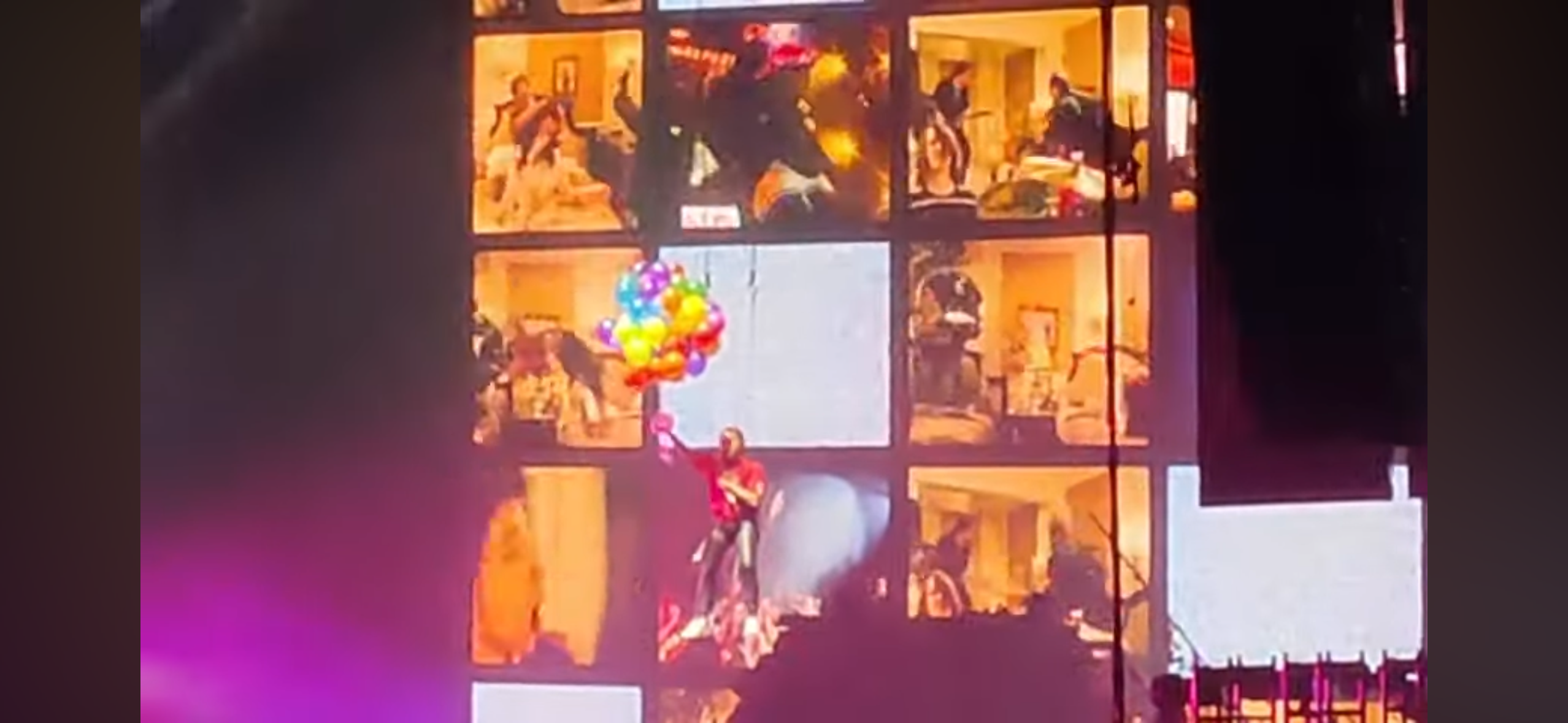 a performer wearing red clothing is holding a bunch of balloons and hangs from a rope during a music concert performance. the stage is lit up with bright lights, and the audience is cheering. the performer is visible through a series of video screens which show the band and audience.