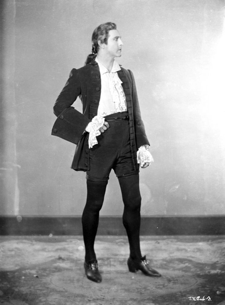 a fulllength black and white image depicts a man in a baroquestyle costume. he is standing with his right hand on his hip and looking to his right. he is wearing a darkcolored coat, a ruffled white shirt, dark breeches, tights, and shoes with small heels. his hair is styled with a slight wave and pulled back into a low ponytail. the background is a plain studio backdrop. the overall aesthetic is vintage and theatrical.
