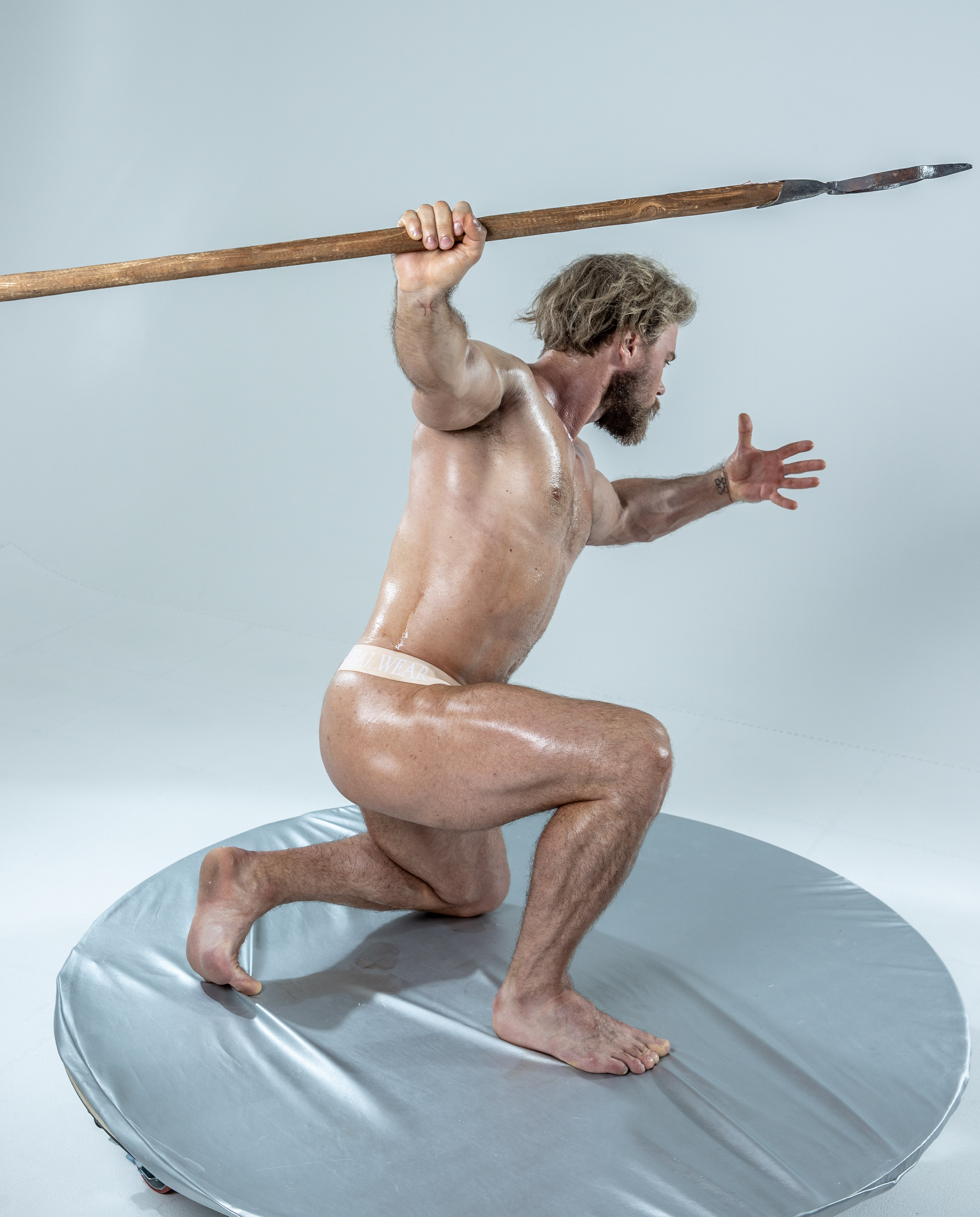 a muscular man with his body oiled up is crouched on a large silver disc, about to launch into action.  he is holding a spear in his right hand, and his body is turned to the right of the image, away from the viewer. the background is a solid gray.