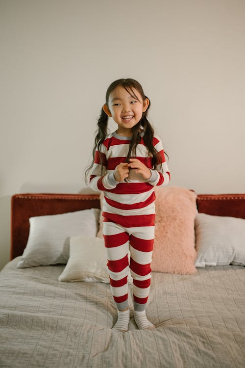 a young girl, approximately 35 years old, is joyfully standing on a bed. she is wearing red and white striped pajamas, which are longsleeved and have a relaxed fit. her dark hair is pulled back into loose pigtails. she is smiling and appears happy and playful, possibly in a home environment, suggesting a casual or relaxed atmosphere. the bed has a plush feel, with pillows and a throw suggesting comfort. the overall tone is cheerful and inviting.