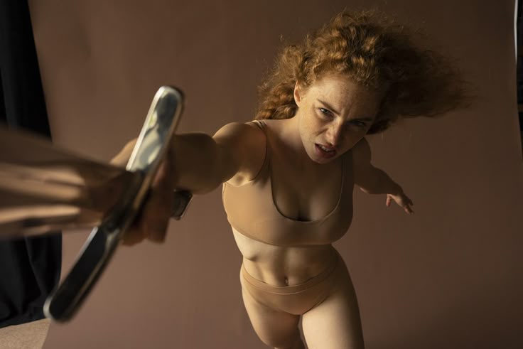 a woman with red hair and freckles is holding a sword in a studio setting. she is wearing tan lingerie and has an angry expression. she is in an action pose, lunging forward with the sword.