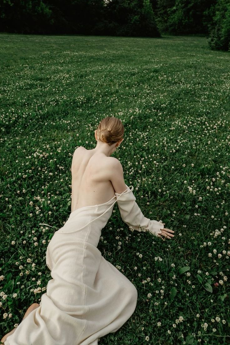 a woman wearing a white, openback dress kneels in a field of clover. she is facing away from the camera. the field is green with small white clover flowers. in the background, there are trees. the woman has her hair up in a bun.
