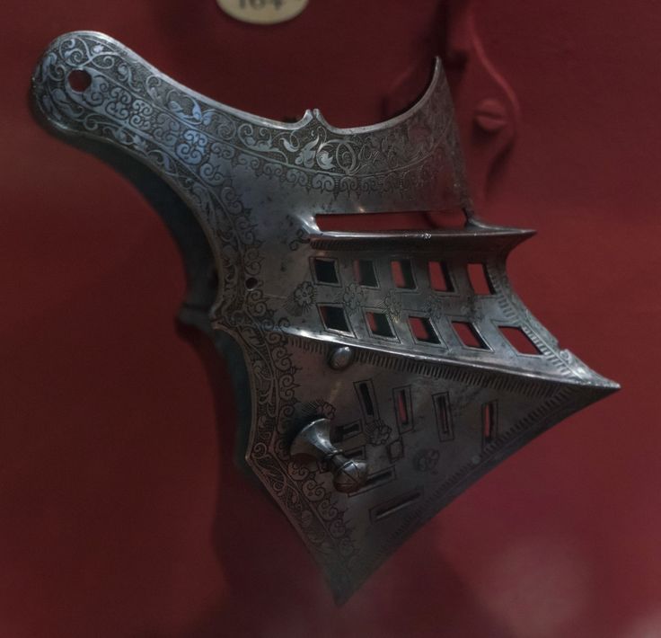 this is a closeup image of a piece of medieval armor, specifically a gorget. the gorget is made of polished steel and is intricately etched with decorative patterns. it is displayed against a deep red background, likely in a museum. the lighting highlights the metallic sheen and the detailed craftsmanship of the armor.