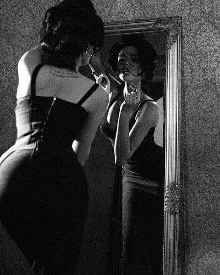 a black and white, closeup shot of a woman applying lipstick in front of an ornate mirror. she's wearing a sleek, dark dress and her reflection is visible in the mirror, creating a symmetrical composition. the image has a vintage, glamorous feel with dramatic lighting and shadows. the background is a patterned wallpaper, further enhancing the retro aesthetic. the woman's focus on her makeup and her expression create a sense of mystery and allure.