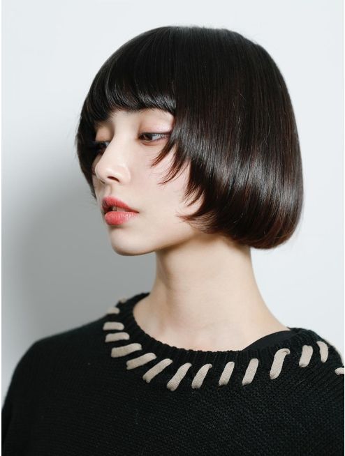 a young woman with short black hair in a bob style, with bangs that cover her forehead, is looking away from the camera. she is wearing a black sweater with a white trim around the neckline. the background is white.