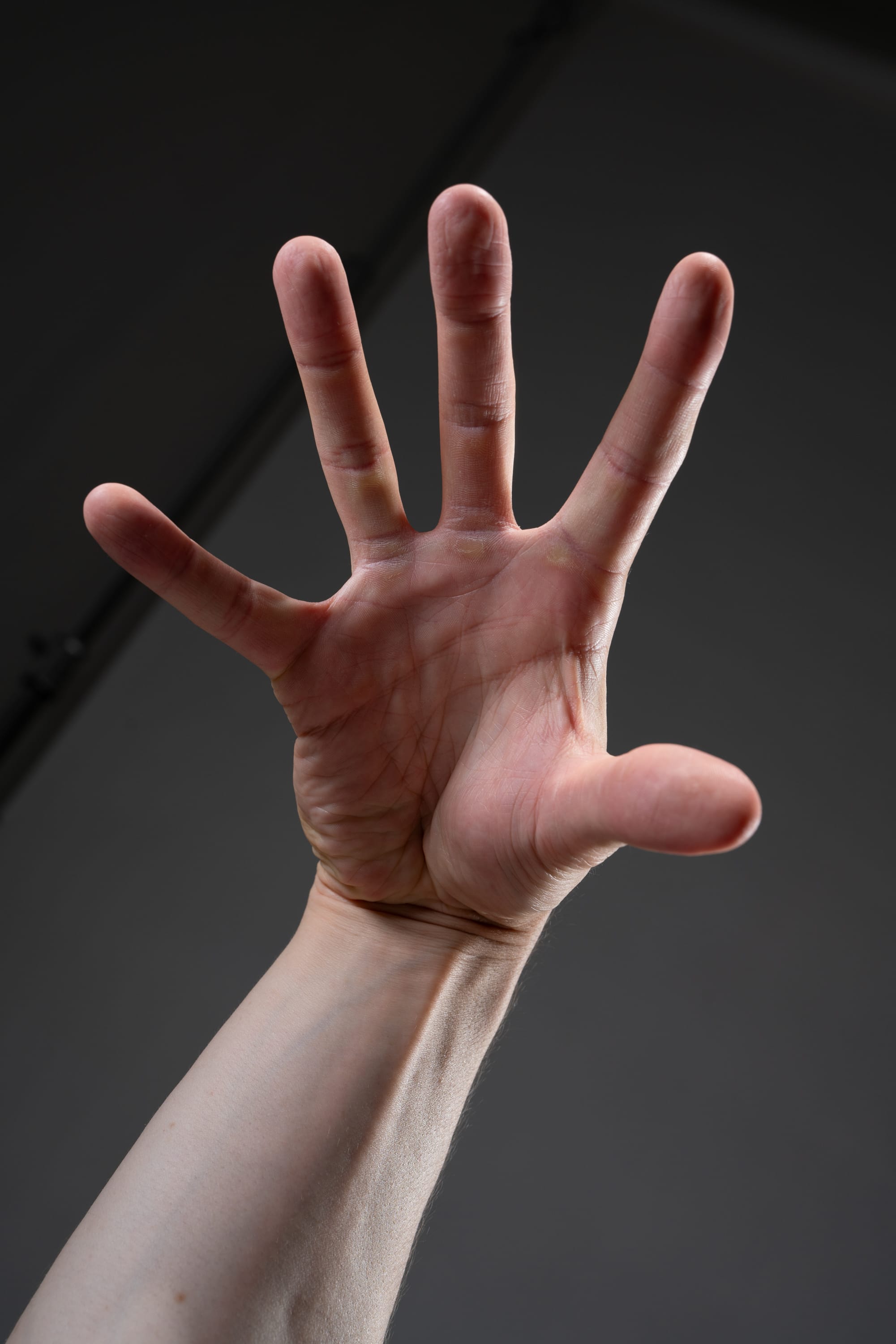 the image shows a person's left hand reaching upward, with all fingers extended. the hand is illuminated by a light source from above. the background is a dark gray color. the image is taken from a low angle view, making the hand appear larger than life.
