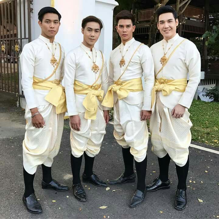 four young men are dressed in elegant white traditional thai clothing. they are standing outdoors, smiling and looking at the camera. they are wearing gold sashes, gold jewelry, and black shoes. they are all dressed in the same style of traditional thai dress. they are standing in a group, looking like they are having fun.