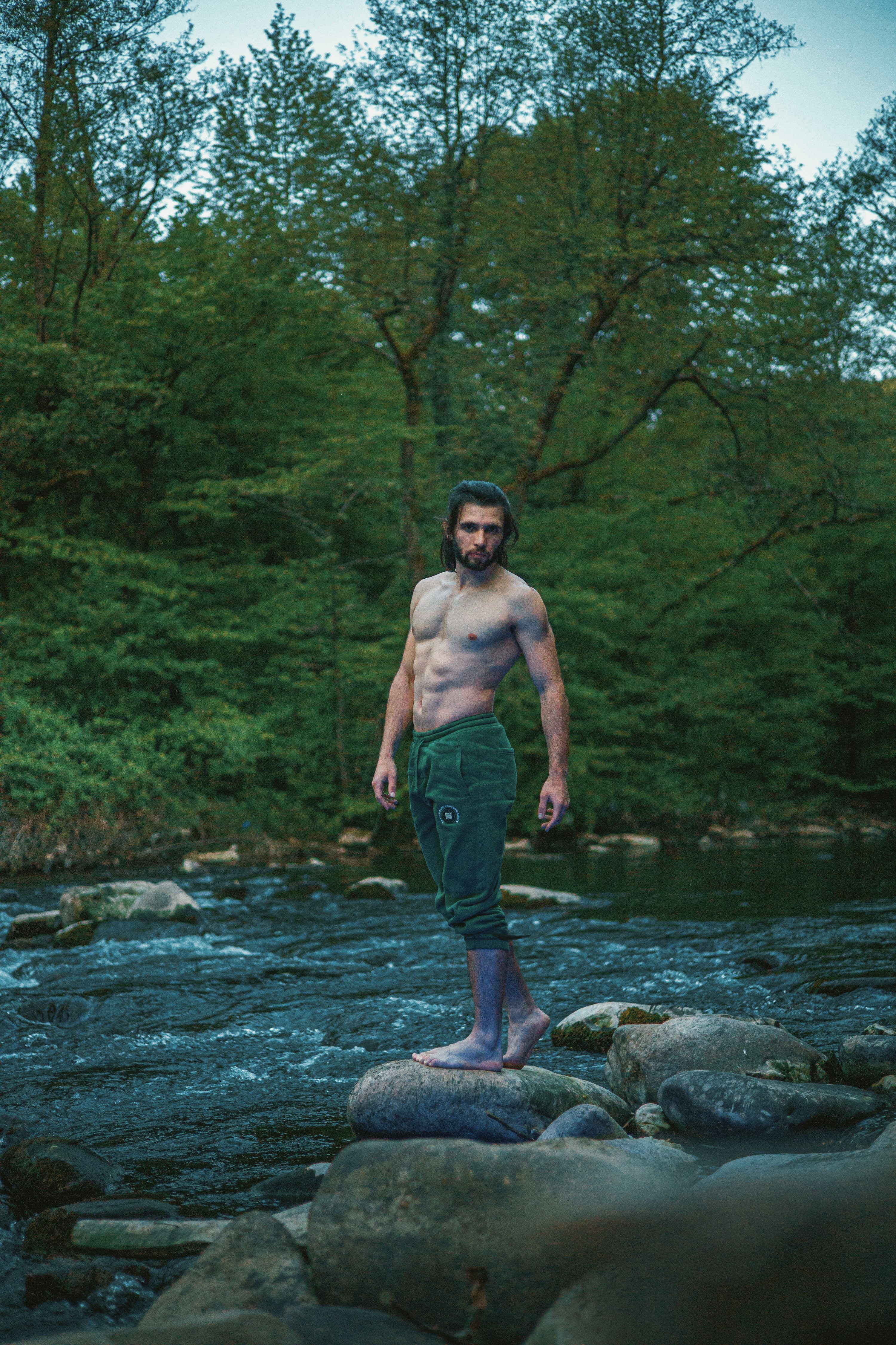 the image shows a muscular, shirtless man with dark hair and a beard standing barefoot on rocks in a shallow river.  he's wearing dark green pants and appears relaxed, almost meditative. the background features a lush green forest, with trees reflected slightly in the dark, flowing water of the river. the overall mood is calm, tranquil, and natural. the lighting suggests it may be daytime. the man's physique is clearly visible, suggesting a fitness or athletic focus.
