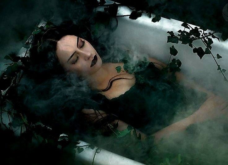 a woman with dark hair and dark clothing lies in a bathtub filled with water.  she has closed eyes and dark lipstick and is surrounded by green plants and smoke.  the image is dark and mysterious, suggesting a gothic or fantasy setting.