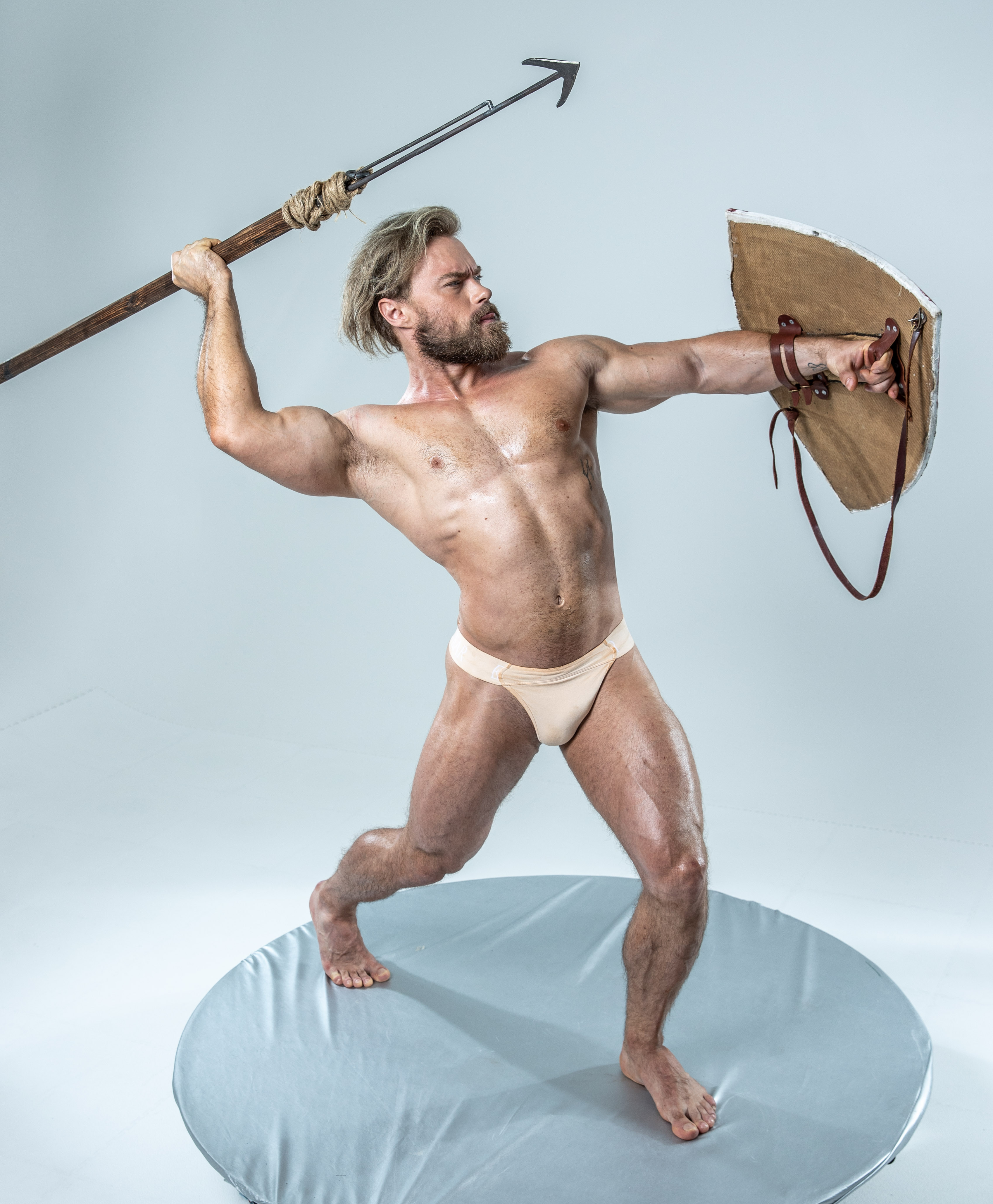 a muscular man with a thick, blonde beard and short hair stands in an attack pose on a gray backdrop in front of a white cyclorama.  he is holding a spear overhead with his right hand and a round shield in his left hand.  the man is wearing a pair of nudecolored briefs.  his body is glistening and oiled and it appears he is in the middle of an action sequence.
