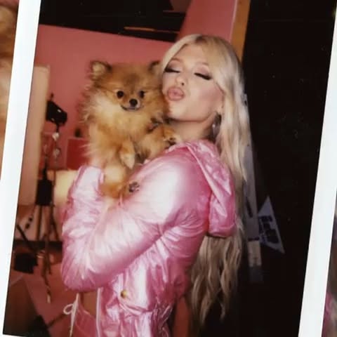 a polaroid photo shows a blonde woman in a pink jacket puckering her lips while holding a pomeranian dog.