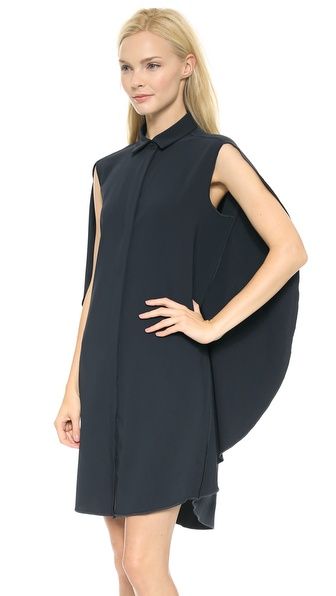 a woman stands in a studio wearing a kneelength, collared, sleeveless dress with a cape detail. she has blonde hair and is facing forward with her right hand on her hip.