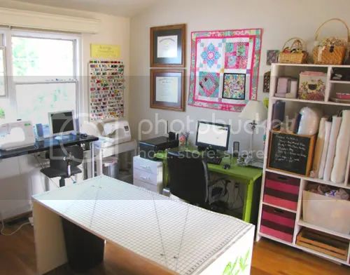 this is a sewing room. there is a sewing machine on the left side of the room and another on the right. the sewing machine on the left is next to a computer. in the middle of the room is a large table with a cutting mat on top. to the right is a green desk with a computer and a chair. a bookshelf is next to the desk. the bookshelf has baskets and other items on it. above the desk there are certificates. there is a quilt on the wall above the desk. there is a window in the upper left part of the room. there are also sewing supplies, a printer, a white board, and other items in the room.