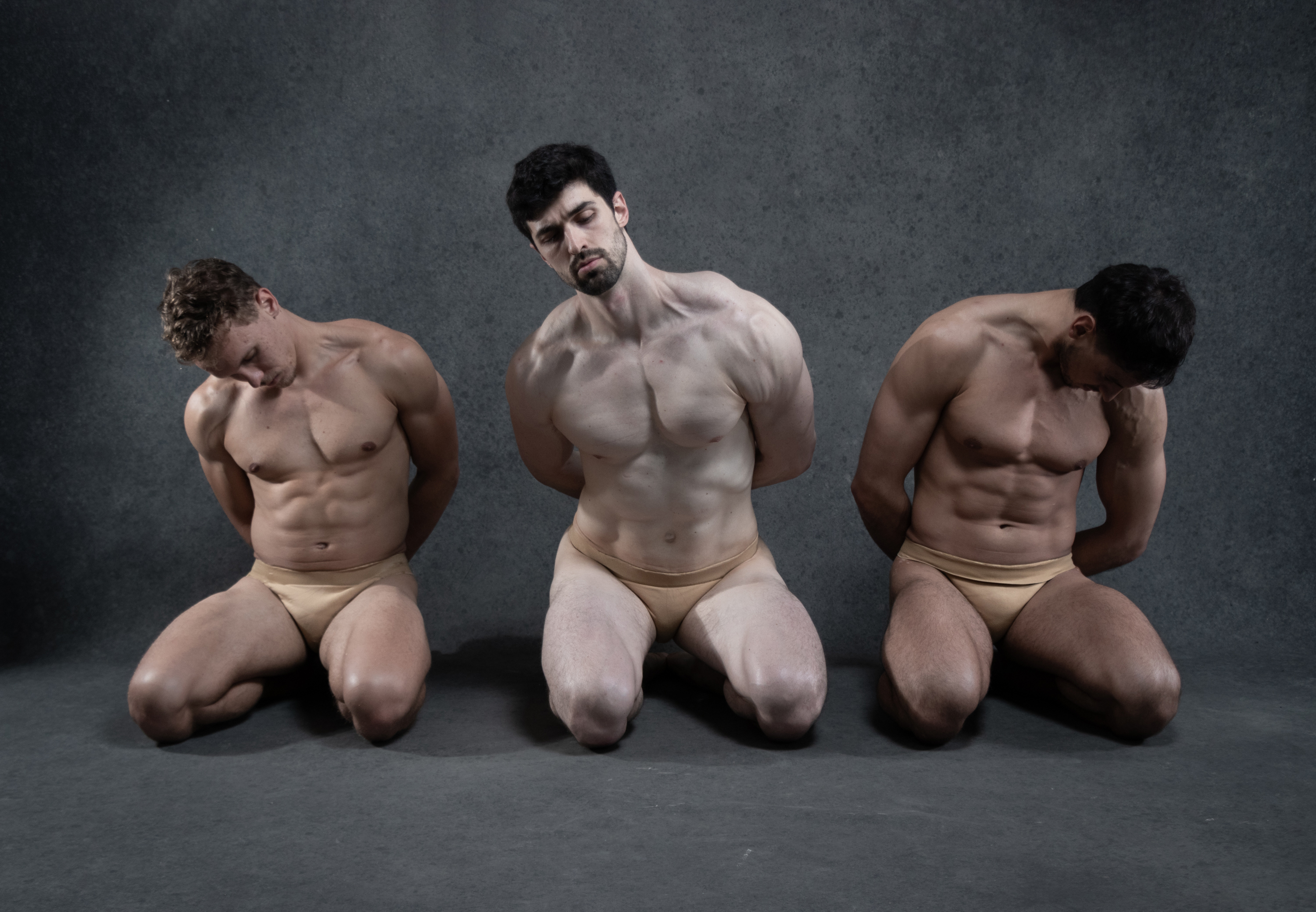 the image captures three muscular male dancers in a studio setting. they are positioned in a line, kneeling with their heads bowed and their arms bound behind their backs. the dancers are shirtless, wearing only skintoned briefs. the lighting is soft yet dramatic, highlighting the contours of their bodies and emphasizing the rawness of their poses. the overall composition evokes a sense of both strength and vulnerability, hinting at themes of confinement and resilience.