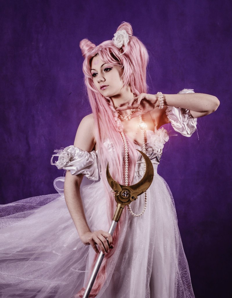 a woman is cosplaying as chibiusa from sailor moon. she is wearing a white dress, a pink wig with the character's signature hair buns, a pearl necklace and bracelet, and is holding the moon stick. the background is a solid purple color.