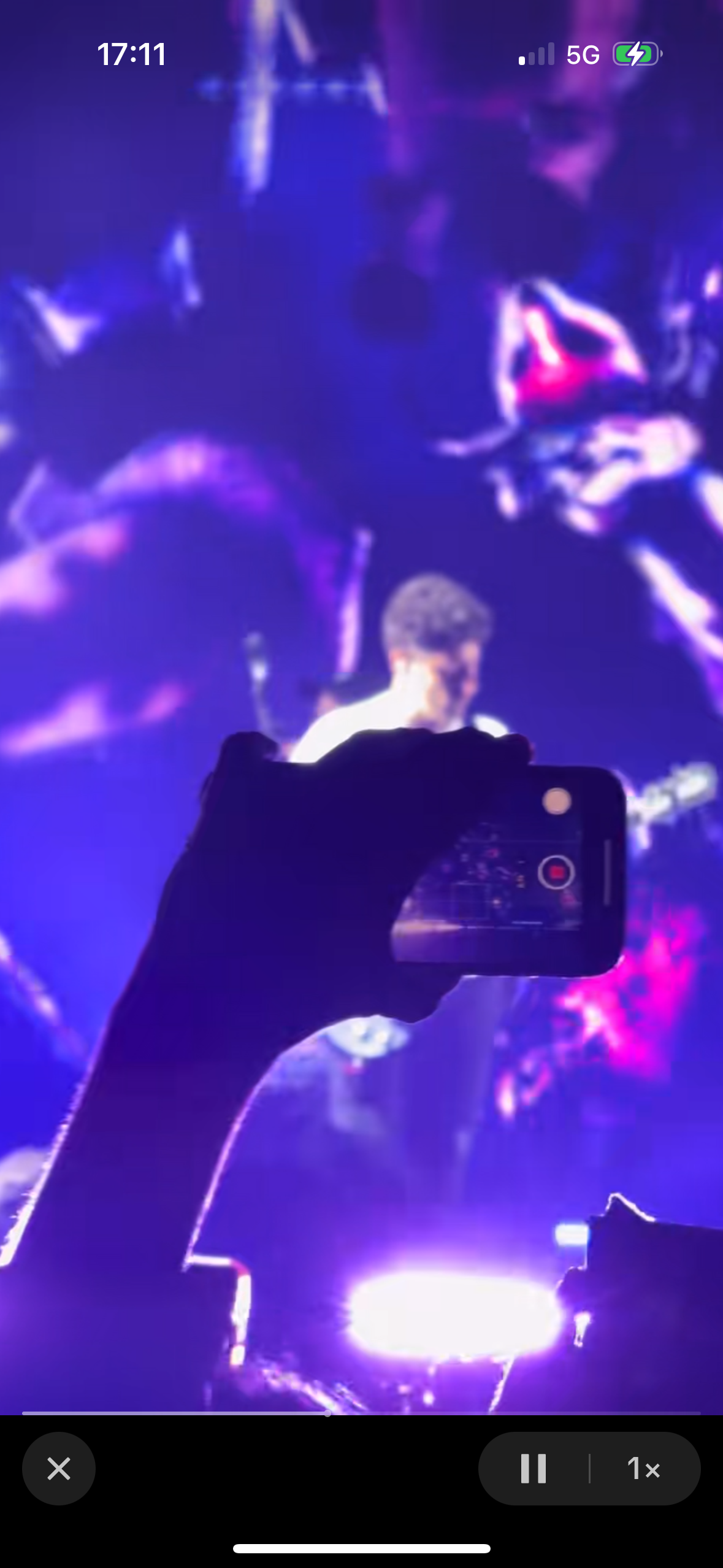 a blurry silhouette of a person filming a concert with their phone. the stage is lit with bright purple lights and the crowd is mostly obscured by the darkness. the person holding the phone is in the foreground, and the stage is in the background. the person in the foreground is capturing a moment at the concert.