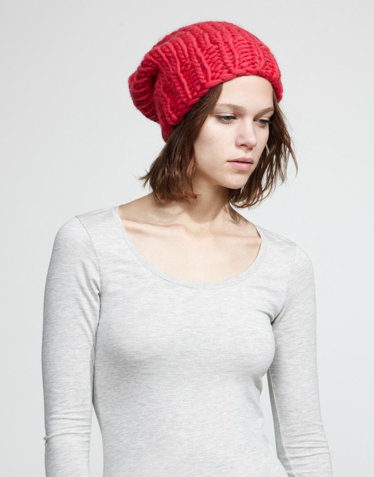 a young woman with short brown hair is wearing a red knit beanie hat. she is also wearing a longsleeved grey top. she is facing the camera and looking down. the background is a light grey.