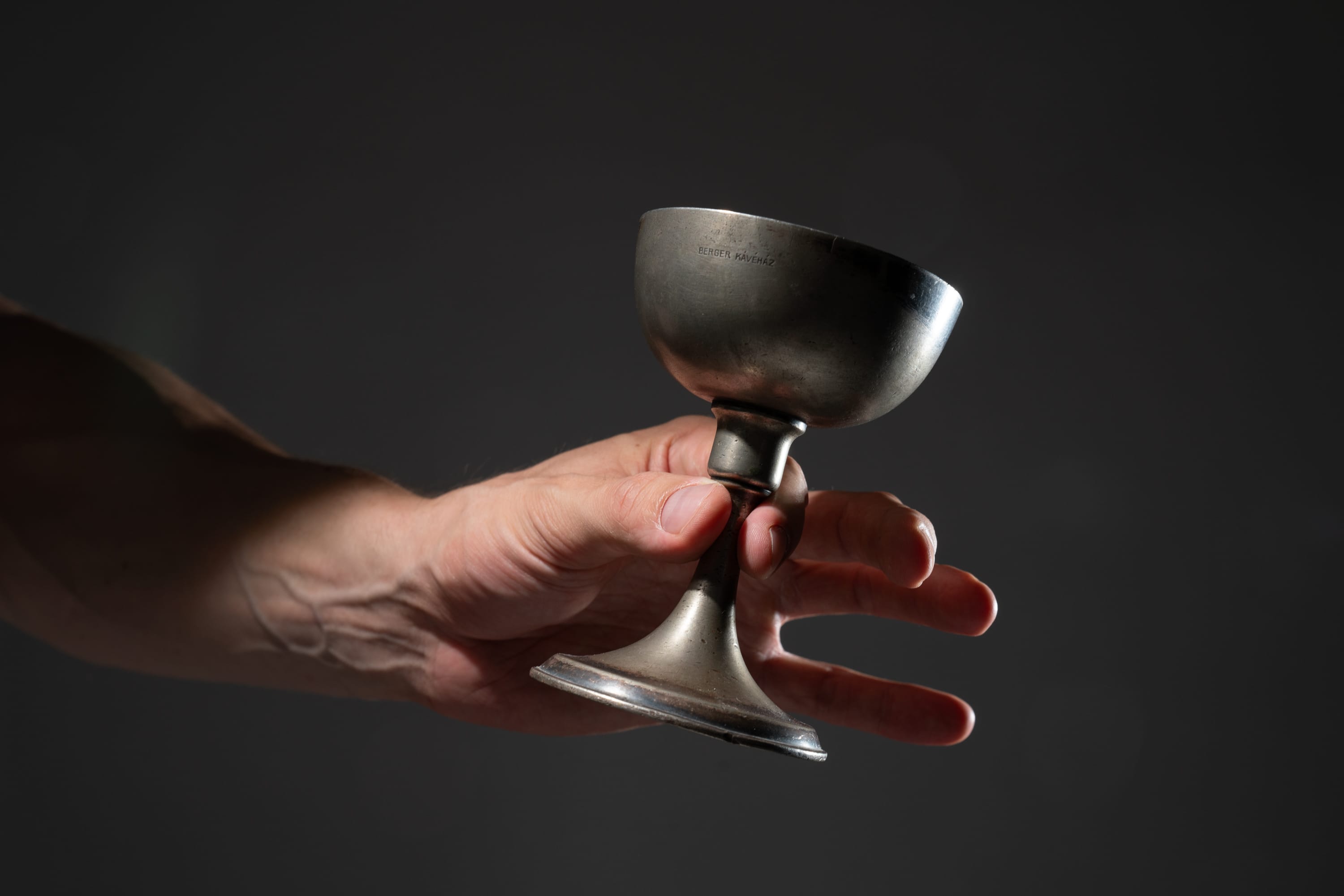 a man holds an antique silver goblet in his hand against a dark background. the goblet is made of tarnished silver and has a rounded bowl and a slender stem. the man's hand is outstretched, and his fingers are curled around the stem of the goblet. the background is dark and shadowy, creating a dramatic and mysterious atmosphere. the goblet is illuminated by a soft light that highlights its intricate details and creates a sense of depth and texture.
