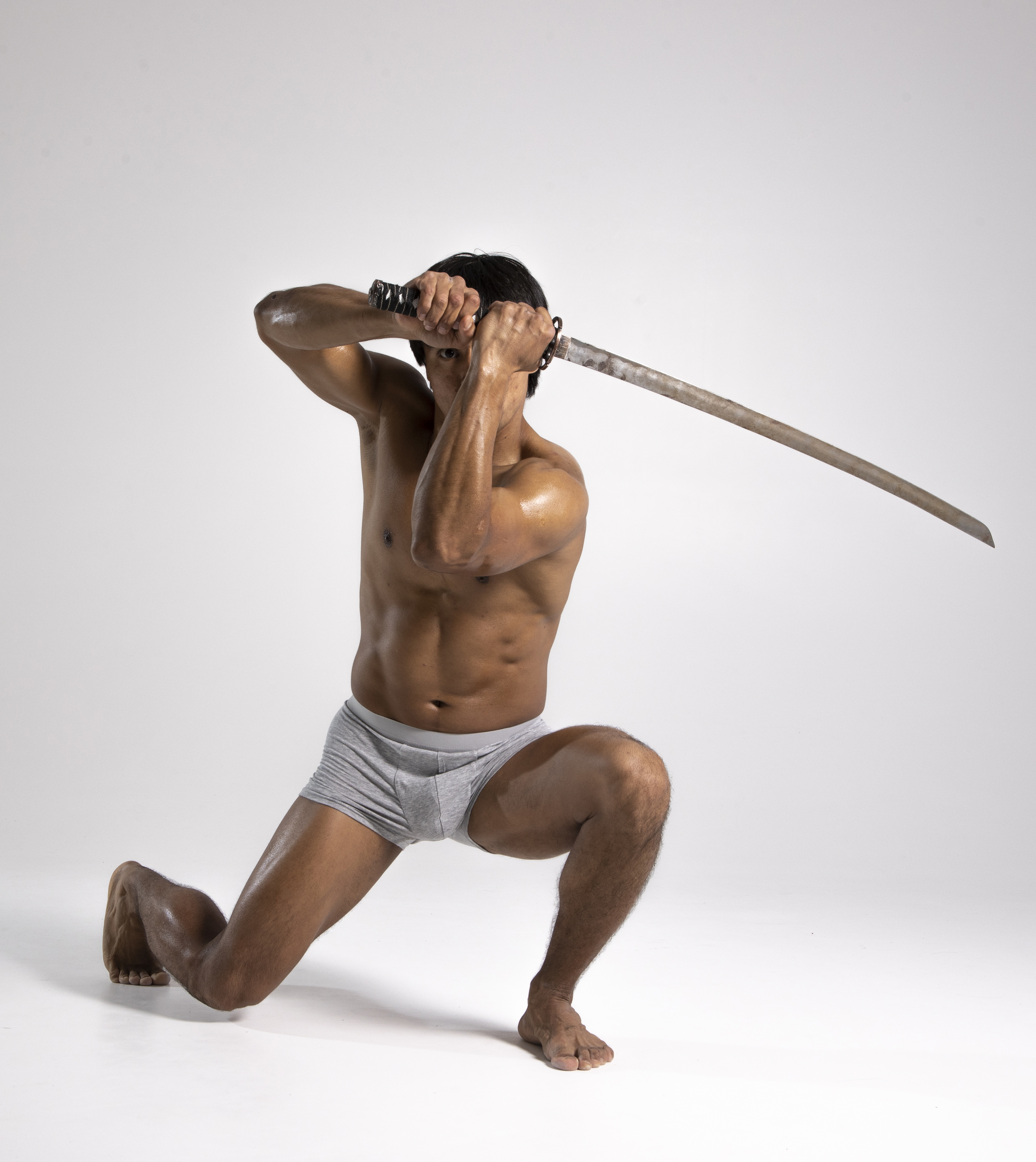 a shirtless, muscular man wearing grey underwear is kneeling in a studio against a grey background, holding a katana sword. he appears to be a warrior or athlete in a defensive or attacking pose.