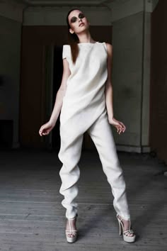 a woman is standing in a studio wearing a white dress and high heels. she is posing for a fashion shoot. her dress is a simple yet elegant design and she is wearing her hair down. her eyes are looking up at the camera.