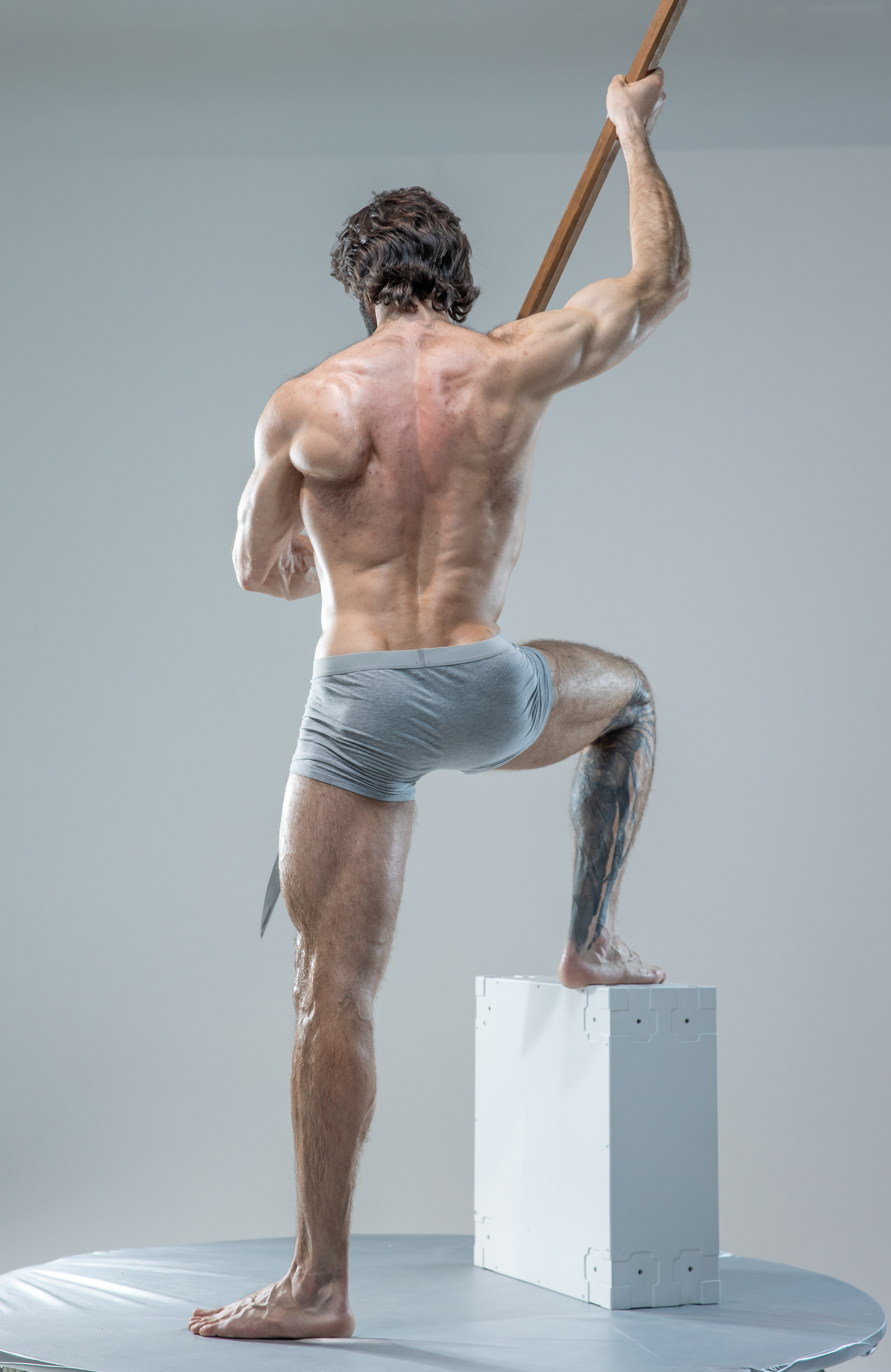 the image shows the back of a muscular man standing on a white platform, holding a wooden spear. he is wearing gray underwear, and his body is turned away from the camera, highlighting his welldefined back muscles. the background is a light gray, and the lighting is soft, creating a natural look. the image has a classic feel, reminiscent of a statue or painting.