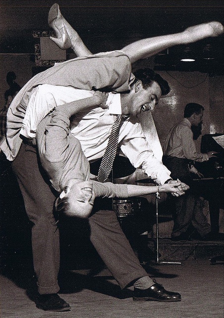 a man and a woman are dancing together in a vintage black and white photo. the man is holding the woman in his arms and they are both smiling. they appear to be dancing the jive or swing. the background shows a man playing the piano and a dark, dimly lit interior space.  the photograph captures a moment of joy and fun.