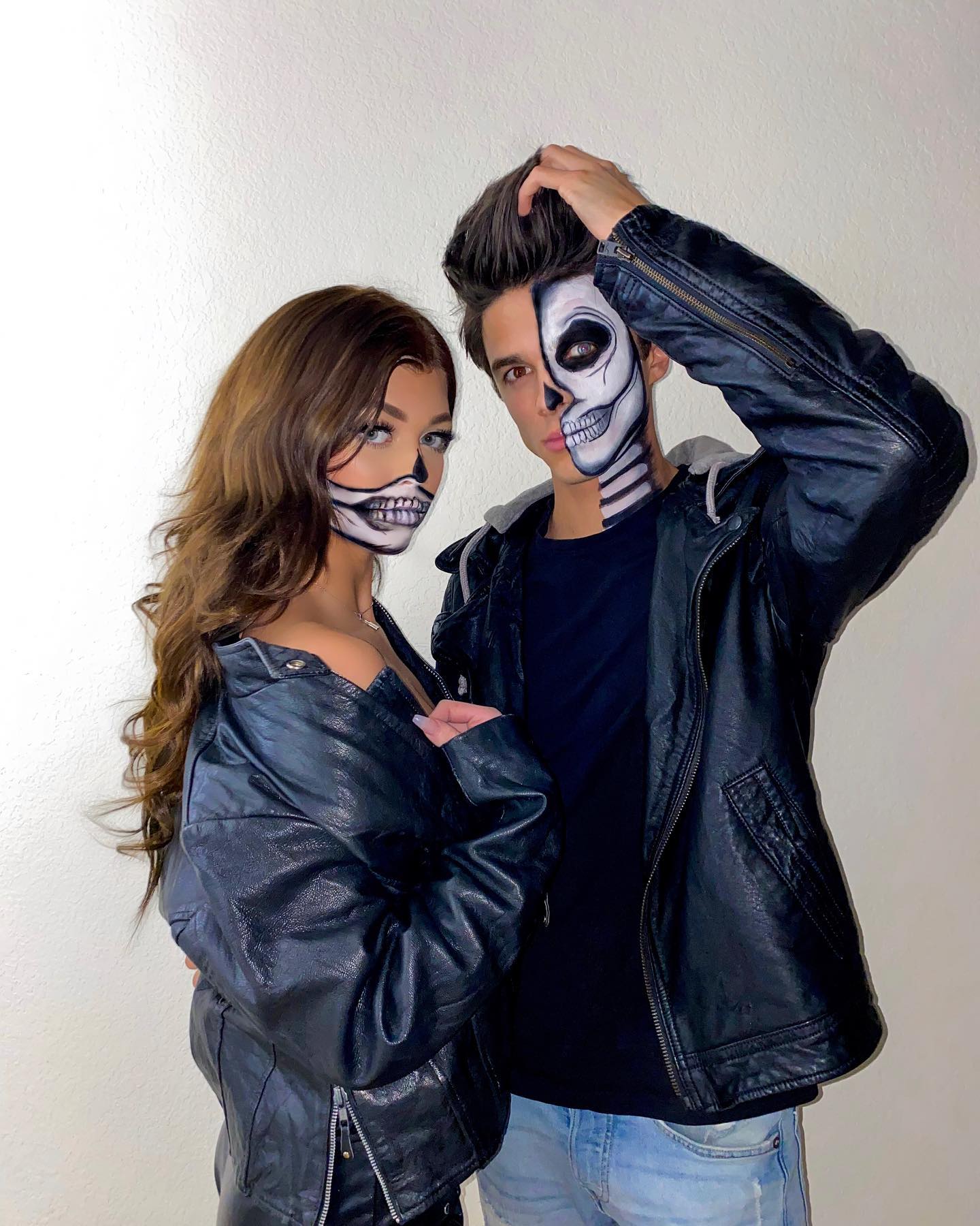 a man and woman dressed up for halloween, both wearing black leather jackets and halfface skeleton makeup. the man is touching his hair, while the woman stands close beside him.