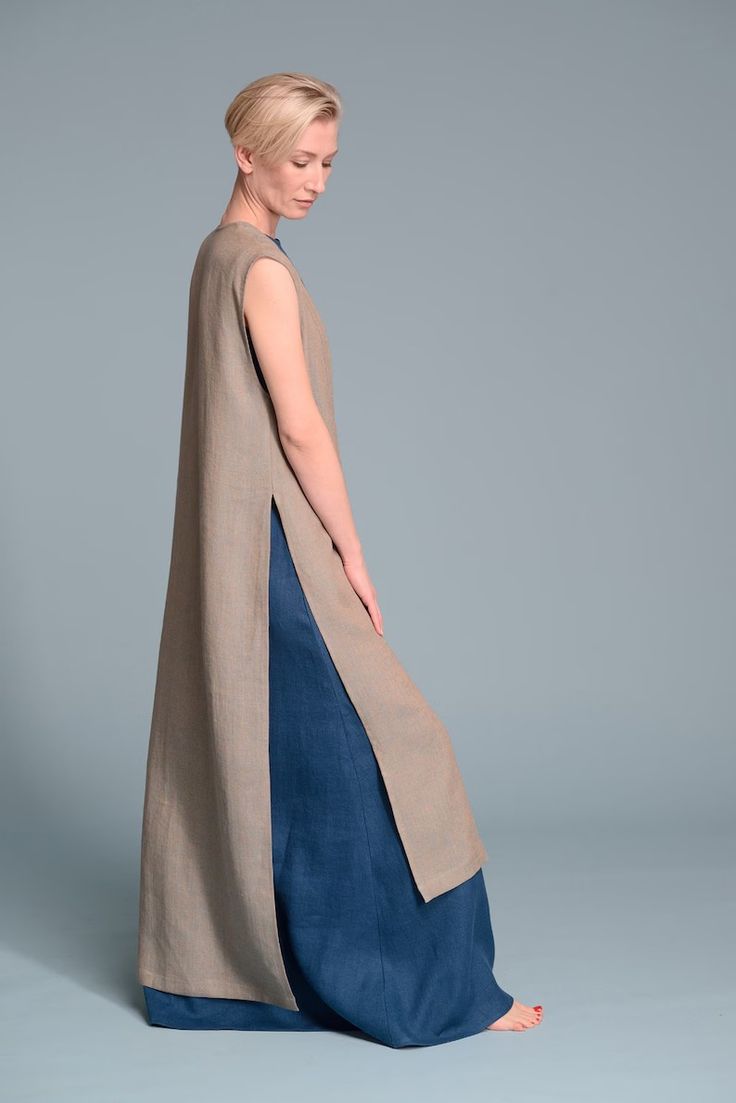 a blonde woman wearing a long grey dress and blue pants is standing and posing in a studio with a grey backdrop. the woman is looking down at her feet and her hair is styled in a short, pixie cut. the dress has a single slit on one side, which reveals the blue pants underneath. the model is wearing no jewelry and her nails are painted red.  the overall look is elegant and casual.