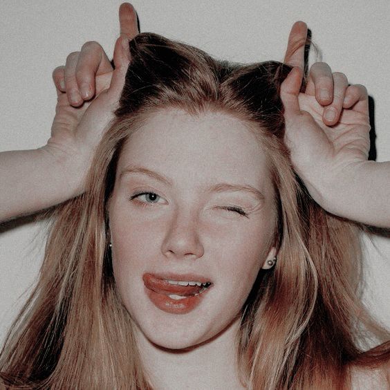 a closeup portrait of a young woman with blonde hair. she's wearing a casual outfit and is making a playful expression by sticking her tongue out and winking. she's surrounded by a light background.