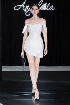 a woman in a white offtheshoulder dress is walking down a runway. she has on silver jewelry and heels. she is looking at the camera as she poses for the photographers.