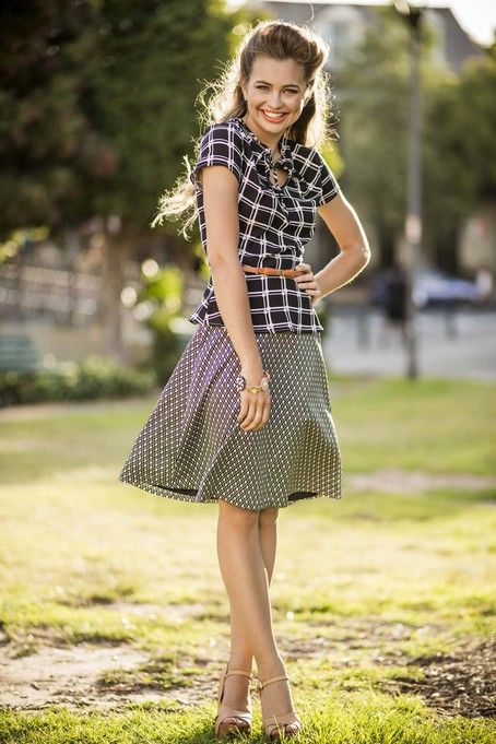 a young woman wearing a plaid shirt and a polka dot skirt is standing in a park. she is smiling and looking at the camera.