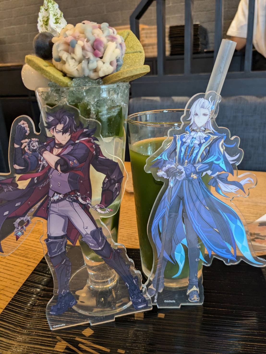 two anime characters stand beside a glass of green tea. they are on a table in a cafe setting. one character has a large, layered dessert topped with whipped cream and a colorful candy in the background. the other character is standing next to the drink.