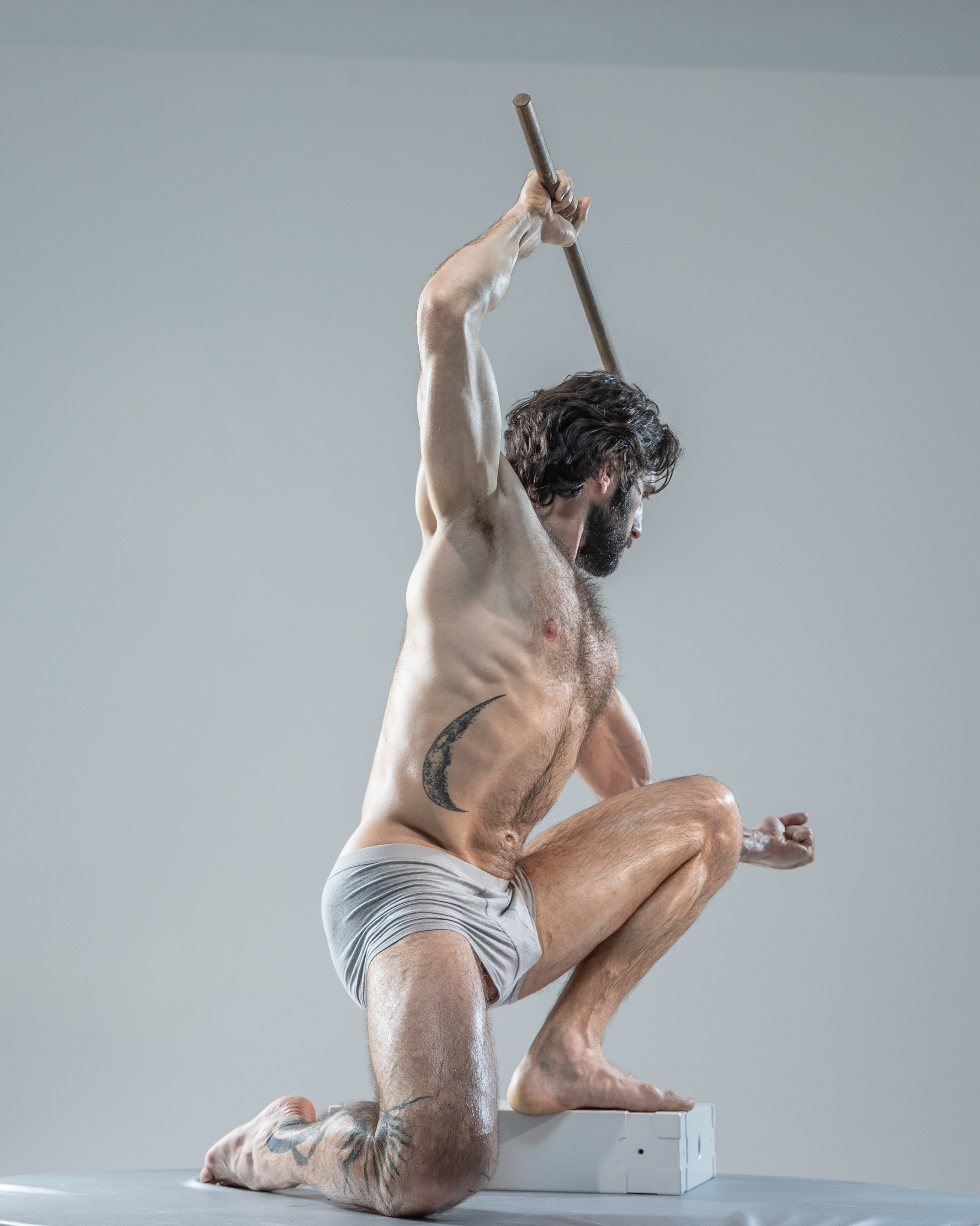 a muscular man with a beard and tattoos is captured midaction in a studio setting. he's shirtless, wearing only grey underwear, and holds a wooden stick above his head as if preparing to strike. his pose is dynamic and powerful, highlighting his physique. the lighting is soft and diffused, creating a sense of focus on his form.