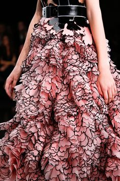 a closeup shot of a model showcasing a stunning pink dress with intricate black ruffle detailing. the dress is a masterpiece of feminine design, with delicate layers of ruffles that cascade down to the floor. the model is wearing a black belt with a silver buckle and is posing with an arm extended towards the side. the background is blurred, focusing attention on the dress.