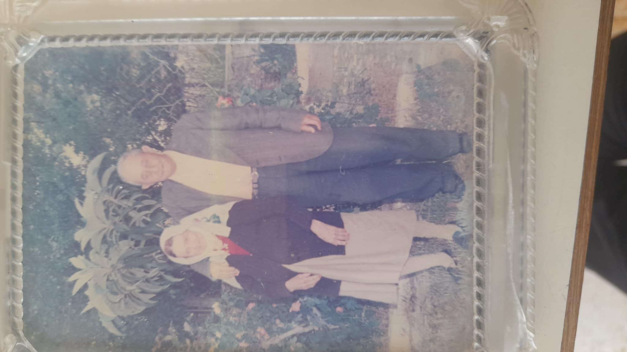the image shows a faded, vintage photograph of an elderly couple posed outdoors in what looks like a garden setting.  the woman is wearing a lightcolored dress and a head covering; the man is in a suit. the background includes lush green plants. the photograph is encased in an ornate, silvercolored frame. the overall style suggests a photo taken sometime in the mid20th century. the image quality is a bit blurry and faded, lending to its nostalgic and vintage appeal.
