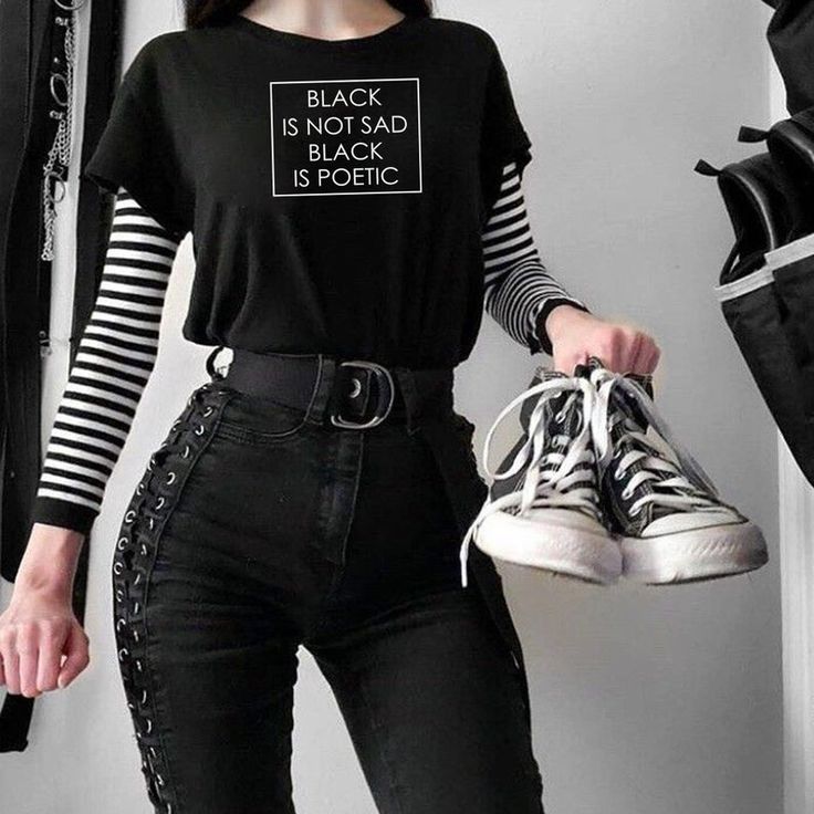 a young woman is wearing a black shirt with "black is not sad, black is poetic" printed on it, paired with a striped longsleeve shirt underneath and high waisted black jeans with laceup detailing down the side. she is holding a pair of black hightop sneakers. the image has a grunge aesthetic, and the woman's style is casual and edgy.